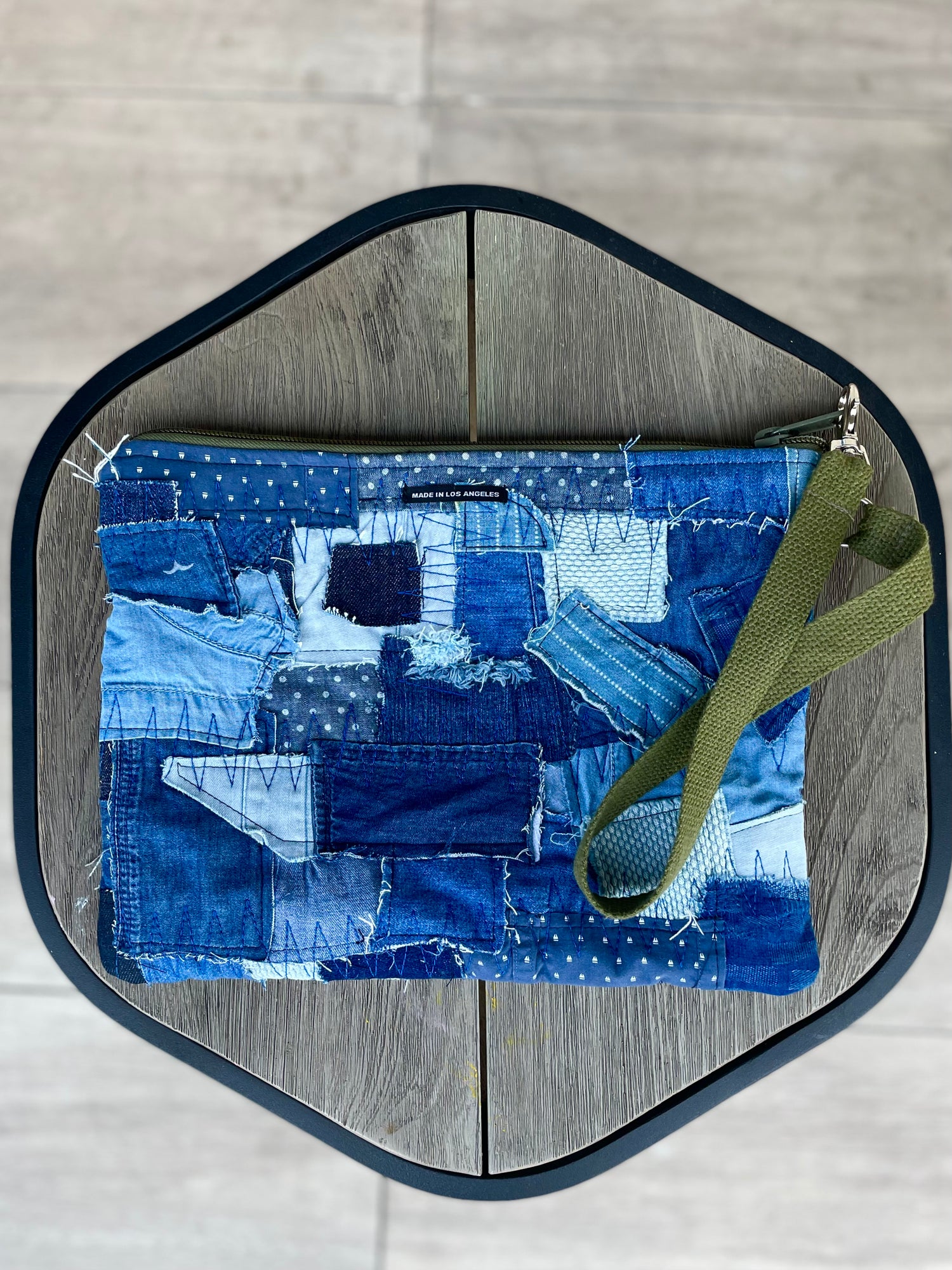 Pouch made from upcycled indigo patchwork denim. Green strap and green zipper. Multifunctional pouch photographed on natural wood stool